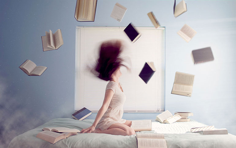 A girl sitting on a bed with books flying around her.