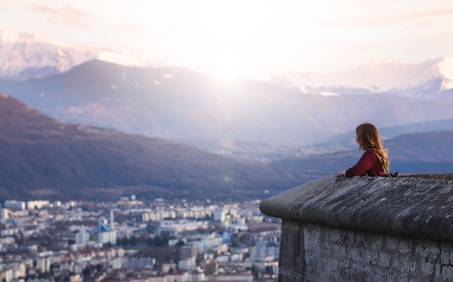 A picture of a person sitting on a ledge overlooking a town and some mountains.
